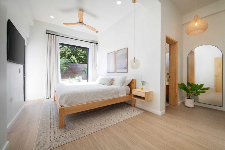 Bright and modern bedroom in a house on Roatan, Bay Islands, Honduras, with white walls, a wooden bed frame, a large window showing lush greenery outside, and minimalistic decor including artwork and pendant lighting.