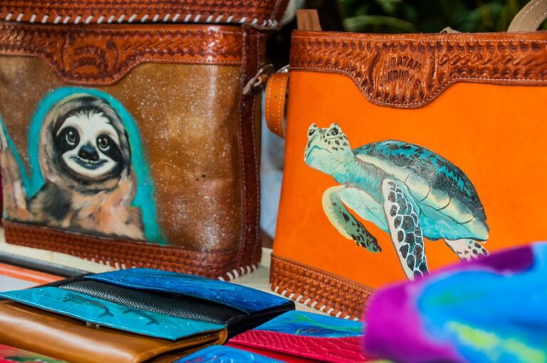 Colorful leather bags featuring animal and jungle-themed artwork are displayed on a shelf in Roatan, Honduras, showcasing local craftsmanship and vibrant designs.