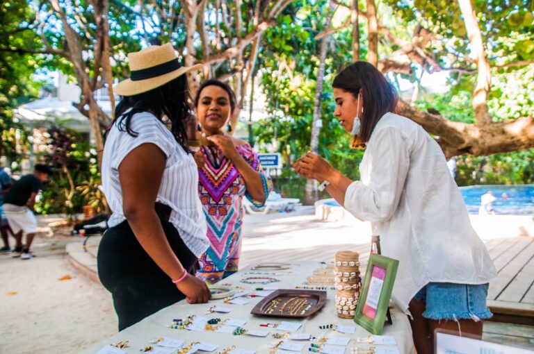 Three women browsing handcrafted jewelry at an outdoor market in Roatan, Honduras, with lush green trees in the background.