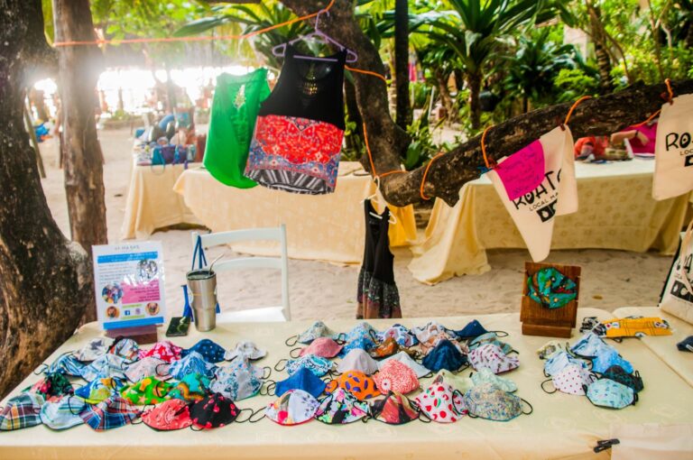 Colorful face masks displayed on a table at an outdoor market in Roatan, Bay Islands, Honduras, with trees and vendor booths in the background.