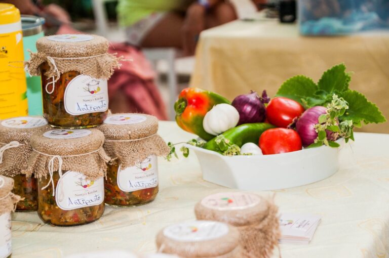 Glass jars with burlap covers labeled 'Antipasto' displayed on a table at a market or food fair in Roatan, Honduras, with fresh vegetables nearby.
