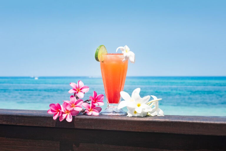 A tropical cocktail garnished with lime and white flowers is displayed on a wooden surface against a backdrop of the clear blue ocean and sky in Roatan, Honduras.