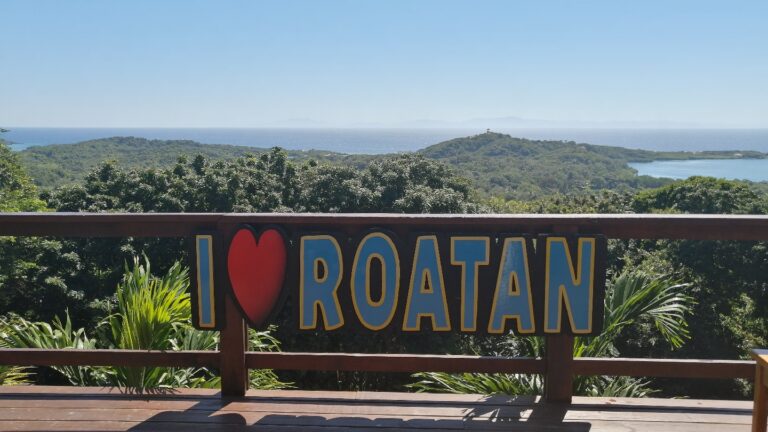 A scenic view of lush green islands and a distant body of water with a lighthouse on a hill, seen from a wooden balcony with a sign reading "I ❤️ Roatan" in bold yellow and blue letters in Honduras.