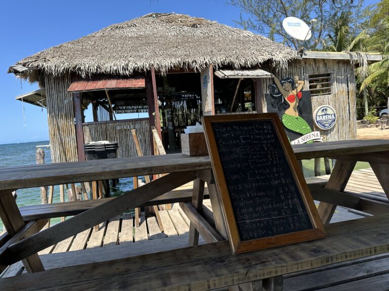 A thatched-roof beachside restaurant or bar on Roatan, Honduras, features a wooden structure with a painted mermaid sign and a chalkboard menu listing various seafood dishes.