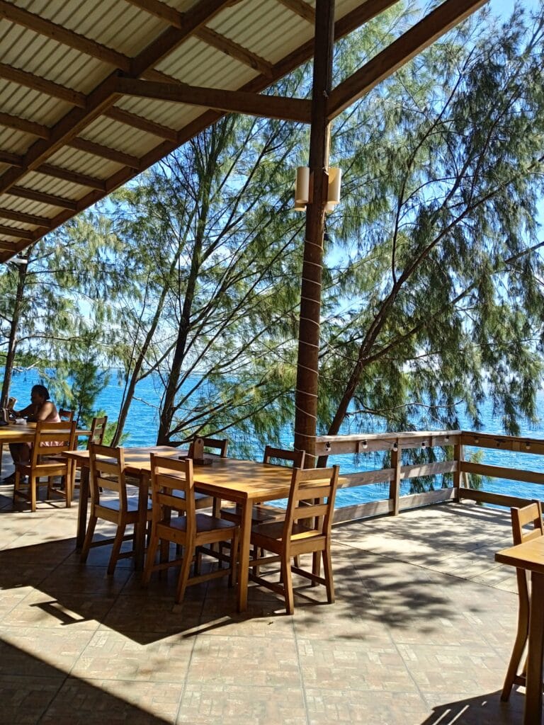 An outdoor dining area in Roatan, Honduras, features wooden tables and chairs on a tiled deck with a view of the ocean, shaded by a roof and surrounded by lush green trees.