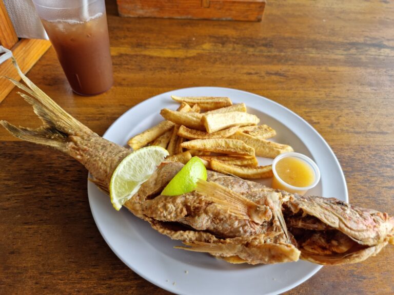 A grilled whole fish served with lime wedges, French fries, and a small cup of dipping sauce on a white plate at a restaurant in Roatan, Honduras.