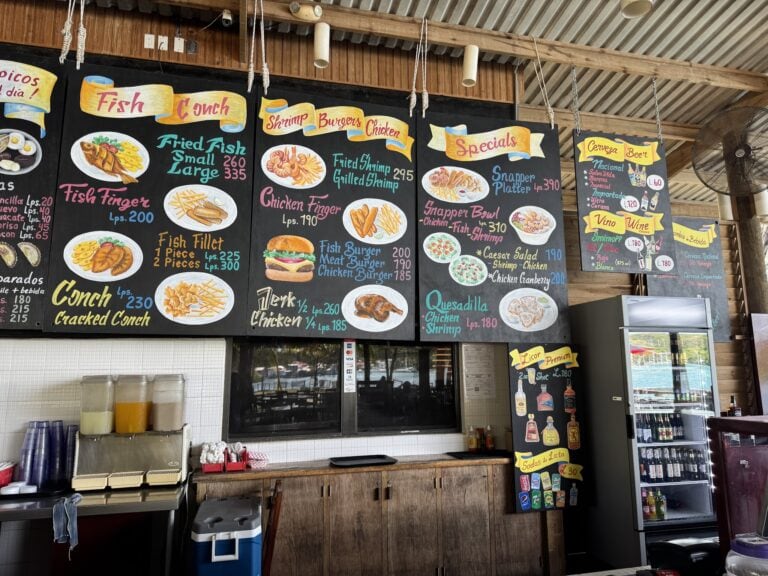 A menu board displaying seafood and drink specials at a restaurant in Roatan, Honduras, featuring items like fried fish, shrimp, chicken, burgers, and various beverages on a black background with colorful handwritten text.