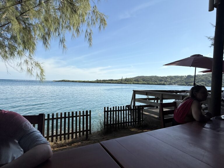 A woman seated at an outdoor dining area on Roatan, Honduras, overlooks a calm body of water with a lush, green hillside and a clear blue sky in the background.