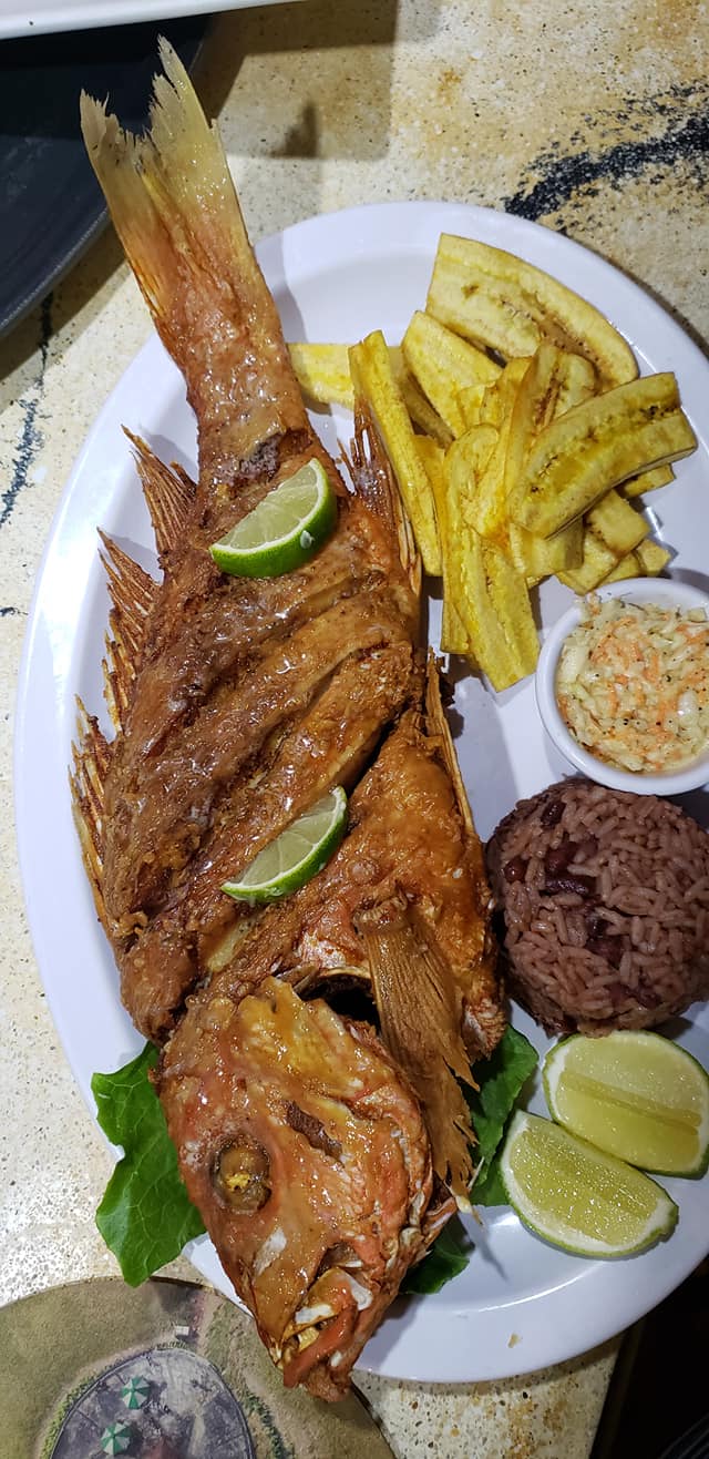 A plate featuring a whole fried fish with lime wedges, French fries, a serving of red rice, and a small cup of coleslaw, set on a white dish at a restaurant in Roatan, Honduras.