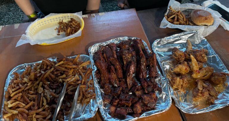 Grilled barbecue ribs, fried chicken wings, and French fries are displayed on aluminum foil and in baskets at a restaurant table in Roatan, Honduras.