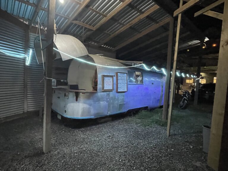 A vintage camper trailer decorated with string lights is parked under a wooden roof structure on a gravel lot in Roatan, Honduras, with a sign advertising onion rings and other menu items.