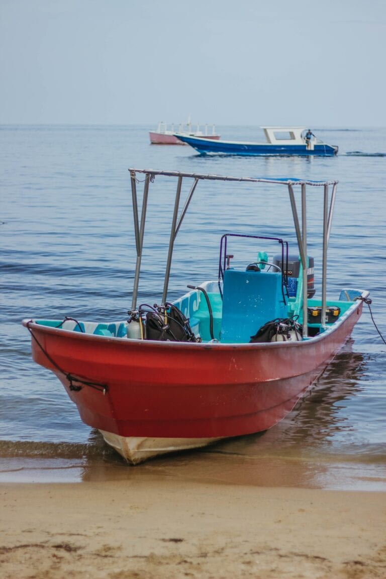 A small red boat with fishing gear on board is anchored near the sandy shoreline in Roatan, Honduras, with two larger boats sailing in the calm ocean in the background.