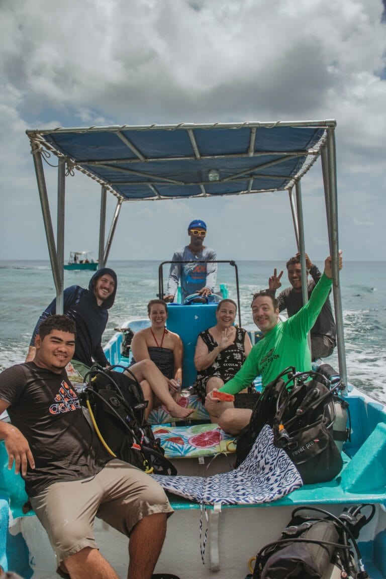 A group of smiling tourists, some with scuba gear, are sitting and standing on a boat with a blue canopy, enjoying a day on the water in Roatan, Honduras.