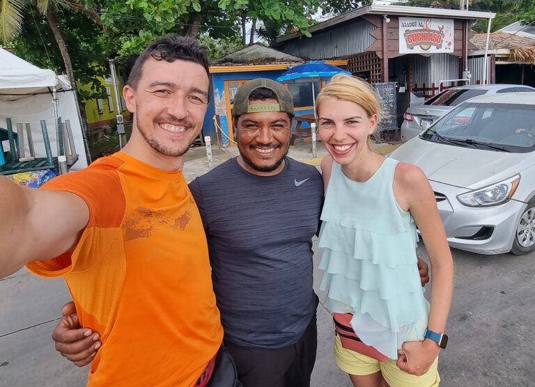 Three smiling people pose together outdoors in front of a restaurant named "Cuchaso" on Roatan, Bay Islands, Honduras, with parked cars, trees, and a signboard visible in the background.