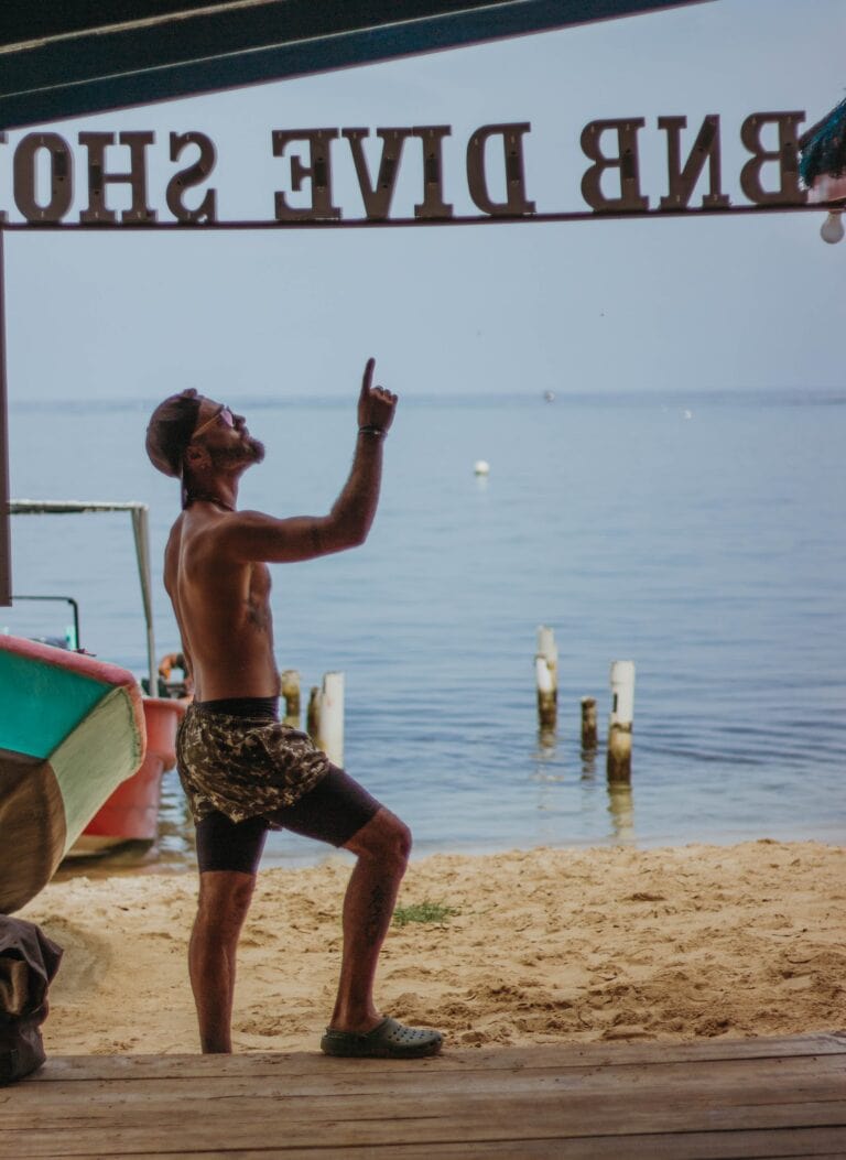 A shirtless man with sunglasses and camouflage shorts is standing on the sandy beach of Roatan, Honduras, pointing upward while near boats and wooden posts by the water.