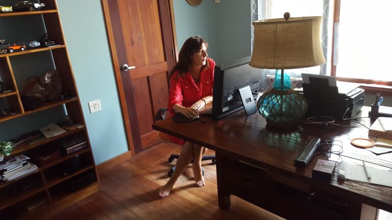 A woman in a red shirt works at a computer desk in a home office on Roatan, Honduras, with a wooden shelf filled with model cars and a large table lamp beside her, near a window with patterned curtains.