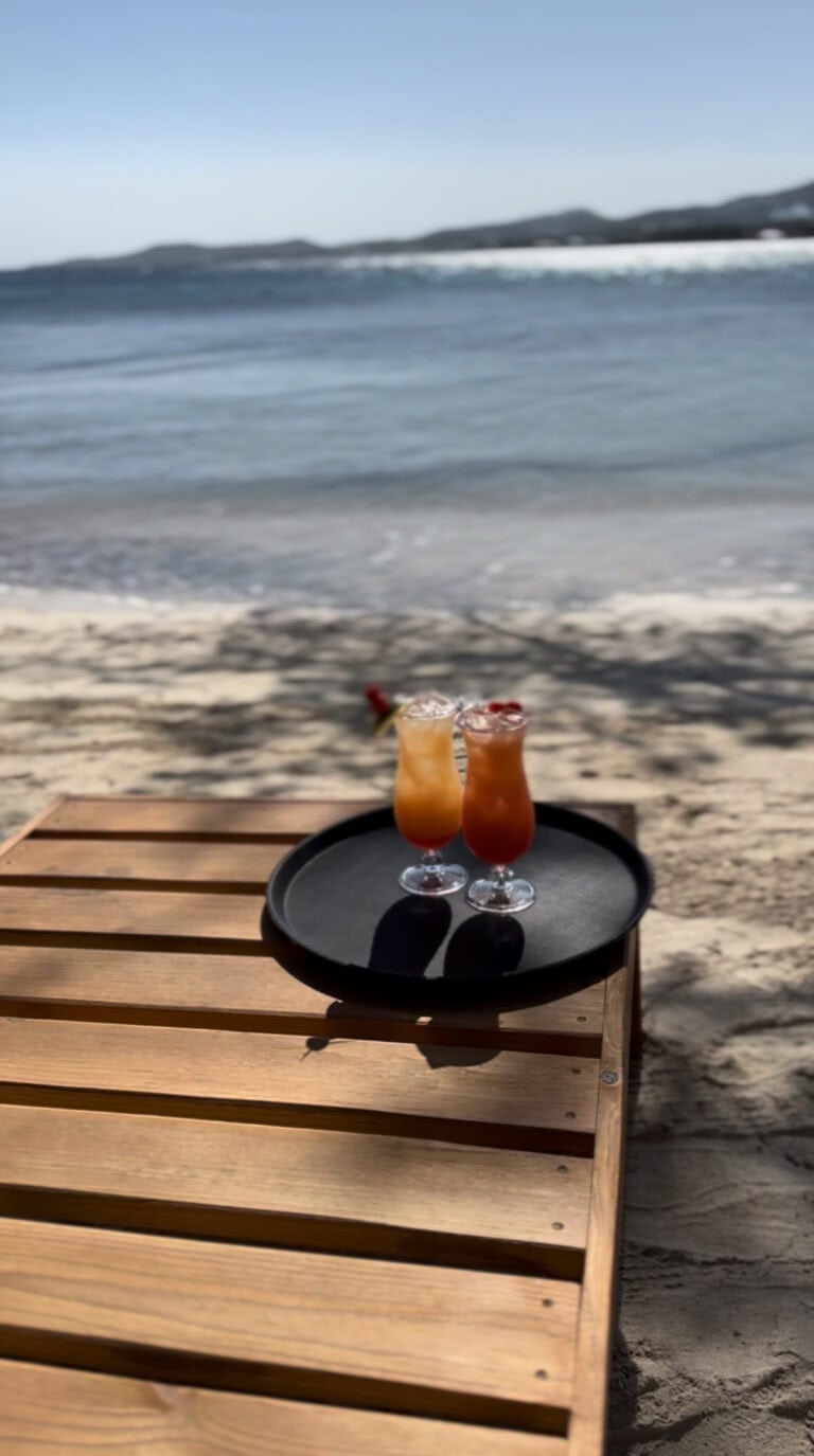 Two tropical cocktails sit on a black tray on a wooden table on the sandy beach in Roatan, Honduras, with the ocean and distant hills visible in the background.