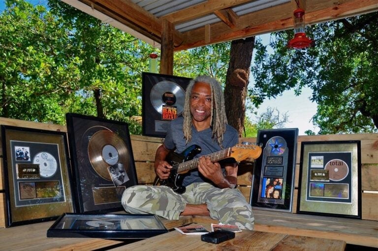 A man with gray dreadlocks plays an electric guitar while surrounded by framed music awards and gold records on a wooden platform open to the outdoors, with green trees and natural light in Roatan, Honduras.