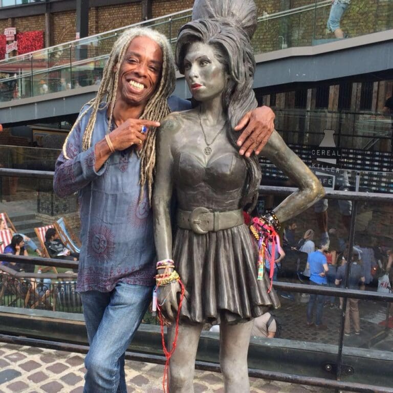 A man with gray dreadlocks is smiling and pointing at a metal sculpture of a woman dressed in a skirt and top, located on a balcony or outdoor terrace in Roatan, Honduras.