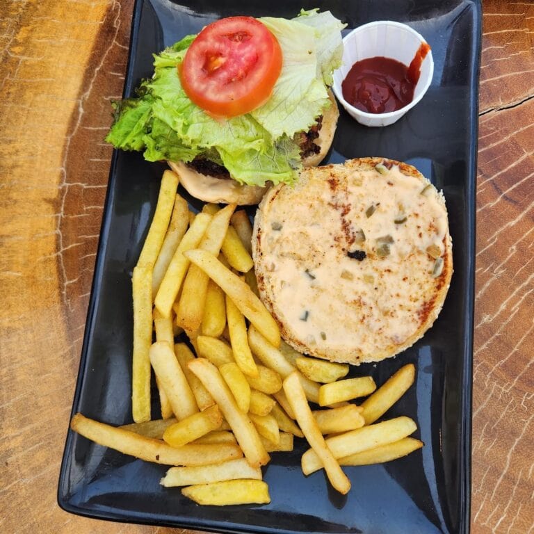 A black rectangular plate holds a cheeseburger topped with lettuce, tomato, and a beef patty, served with a side of French fries and a small cup of ketchup, set on a wooden table in Roatan, Honduras.