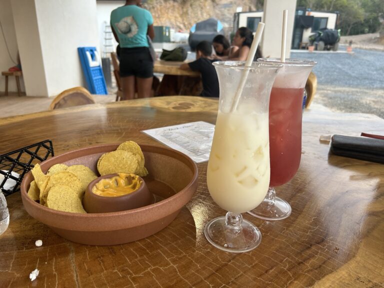 A table at a Roatan restaurant in Honduras features two frosty cocktails—one creamy white and the other reddish—along with a bowl of tortilla chips topped with cheese and salsa, with a group of people sitting at another table in the background.
