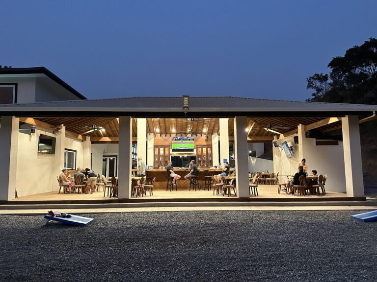 A covered outdoor dining area with wooden tables and chairs, people seated and socializing, a bar with a television showing sports, and a nighttime sky in Roatan, Honduras.