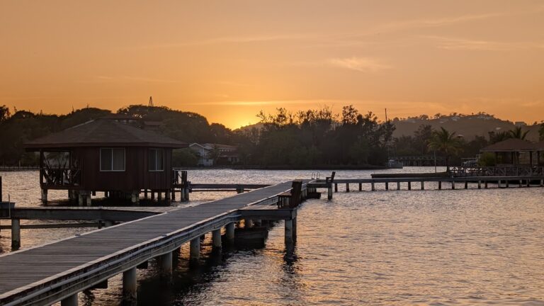 A wooden overwater bungalow with a dock extending over the water on Roatan, Honduras, during a sunset with orange and pink skies and lush green hills in the background.
