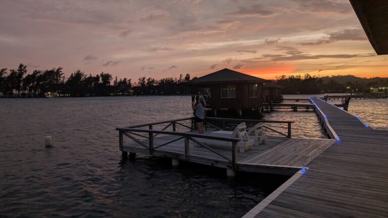A woman stands on a wooden dock overlooking the water at sunset on Roatan, Honduras, with overwater bungalows and a glowing pathway illuminated by blue lights leading to the small structures.