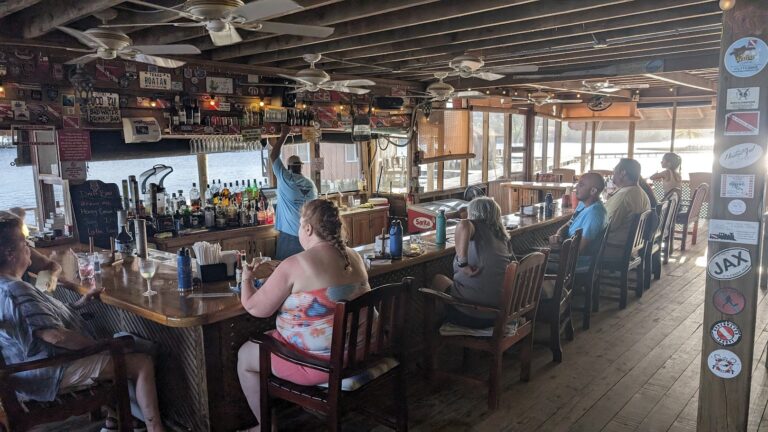 A group of people enjoying drinks at a waterfront bar in Roatan, Honduras, with a view of the water through large windows and tropical decor inside.
