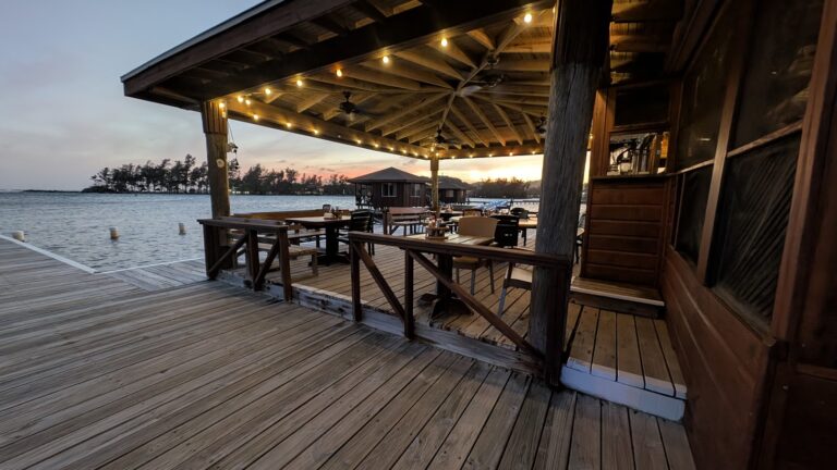 A wooden waterfront restaurant in Roatan, Honduras, features outdoor seating with tables and chairs under a string-lit, open-air roof overlooking the water at sunset.