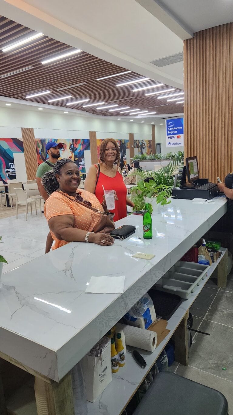 Two women are smiling and sitting at a white marble kitchen counter with a potted plant, while a man stands behind them in a brightly lit indoor space with wood accents and colorful wall art in Roatan, Honduras.