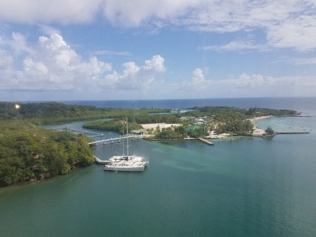 A white sailboat is docked in a narrow, tranquil waterway surrounded by lush green trees, with a view of a beach and the open ocean in Roatan, Honduras, under a partly cloudy sky.