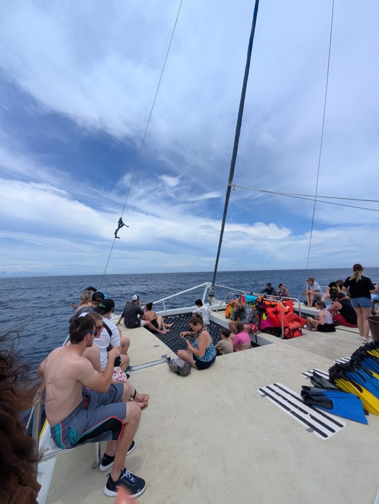 People sitting and relaxing on the pier in Roatan, Honduras, with the sea, sky, and a sailboat mast visible in the background.
