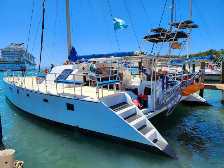 A marina in Roatan, Honduras, features several sailboats docked along the pier with a cruise ship visible in the background under a clear blue sky.