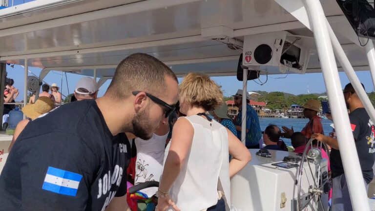 Passengers on a boat in Roatan, Honduras, enjoy a sunny day with clear blue skies and a view of waterfront buildings and greenery in the background.