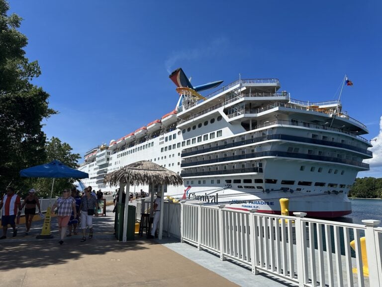 A large cruise ship docked at the Roatan, Honduras port with people walking along the waterfront beneath a thatched parasol and blue sky overhead.