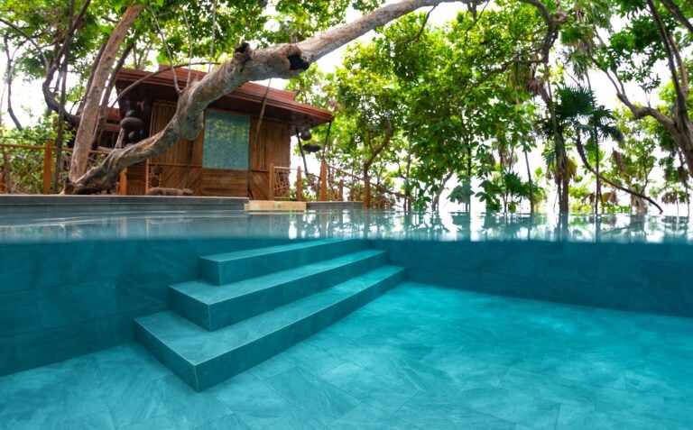 An infinity pool with turquoise water is surrounded by lush green trees and a wooden deck with a small hut in Roatan, Honduras.