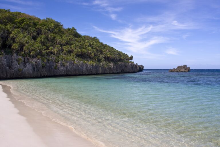A tropical beach in Roatan, Honduras, features clear turquoise waters, white sandy shorelines, a rocky island formation, and lush green hillside with palm trees under a blue sky.