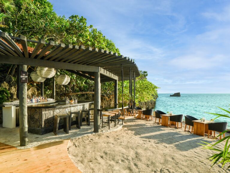 A beachfront bar in Roatan, Honduras, features a wooden deck with hanging lanterns, outdoor seating, and a view of the ocean and a small rocky island in the distance under a clear blue sky.