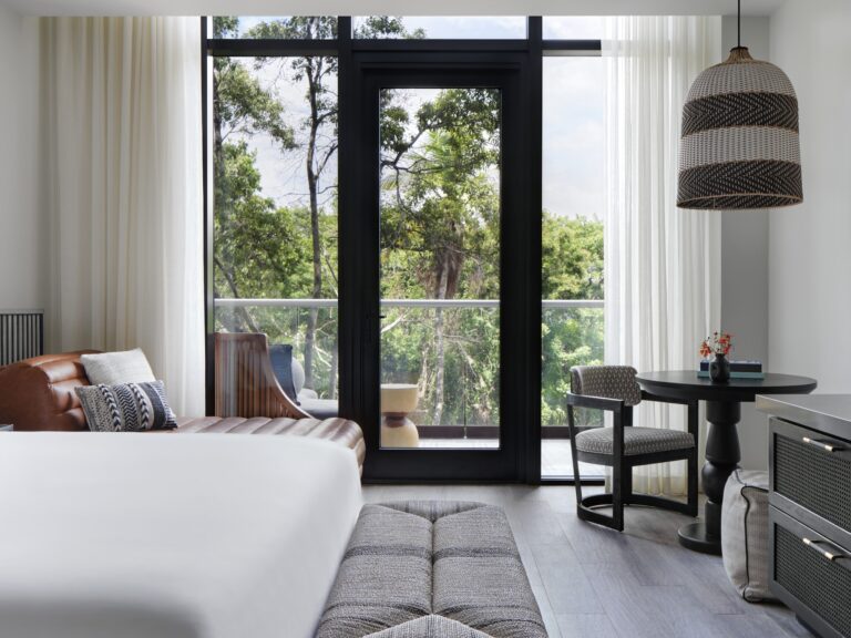 A modern living room in Roatan, Honduras, features large glass doors with sheer white curtains overlooking lush green trees and a balcony, with stylish furniture including a leather sofa with decorative pillows, a round dining table with patterned chairs, and a woven pendant light hanging from the ceiling.