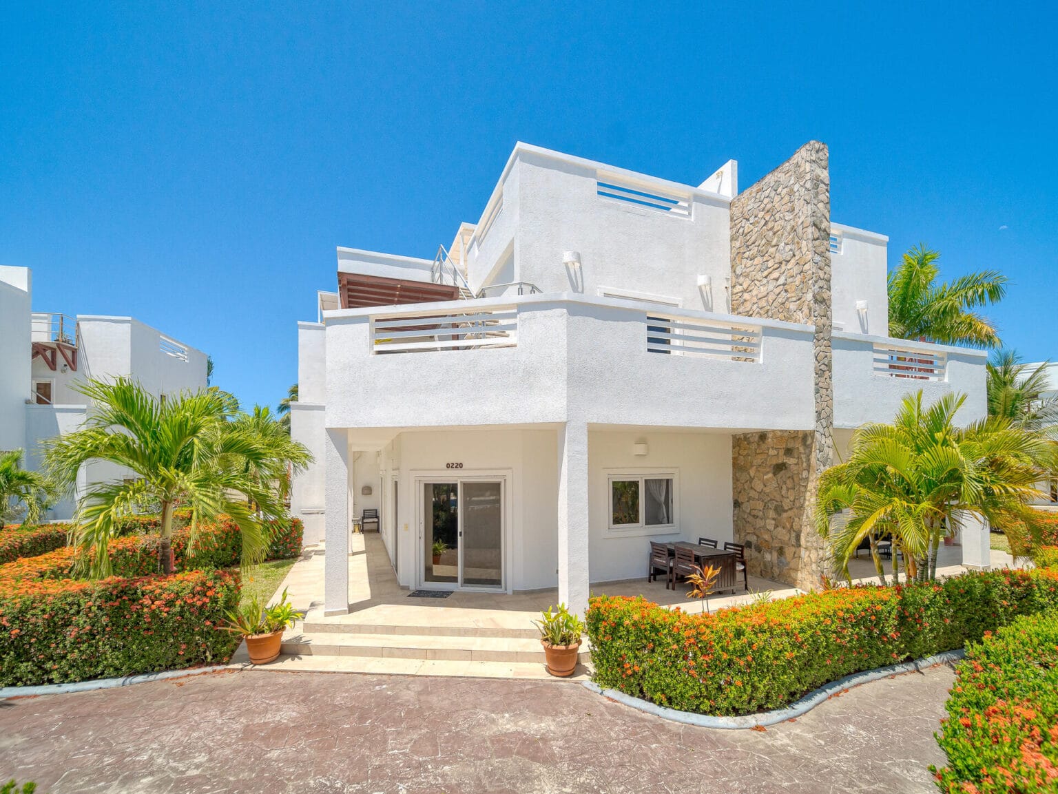 A white, modern three-story building with stone accent walls, surrounded by lush tropical plants and flowering bushes, located in Roatan, Honduras.