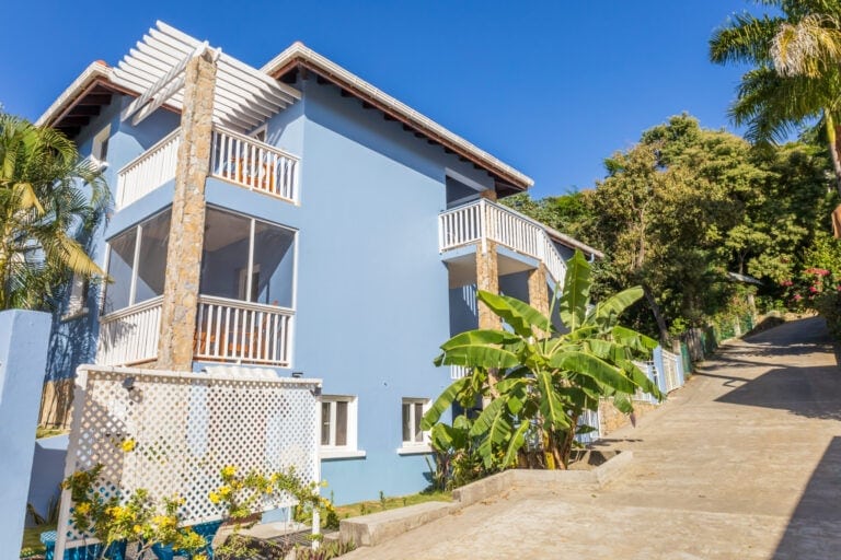 A light blue multi-story building with white railings and stone accents is surrounded by lush tropical plants, including banana trees and flowering shrubs, on a sunny street in Roatan, Honduras.