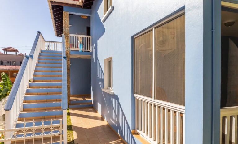 A light blue multi-story building in Roatan, Honduras features an exterior staircase with white railings, leading to a balcony with potted plants, large windows, and a small tiled porch area.
