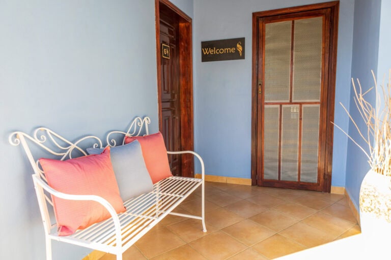 A cozy indoor space in Roatan, Honduras, features a white metal bench with pink and light blue cushions against a blue wall, with a wooden door and a "Welcome" sign with an ice cream cone graphic on the wall.