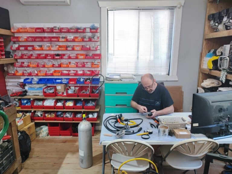 A man working on electronic or technical equipment at a cluttered workbench inside a hardware or electronics shop on Roatan, Honduras, with shelves of small hardware parts, tools, and a window with blinds in the background.