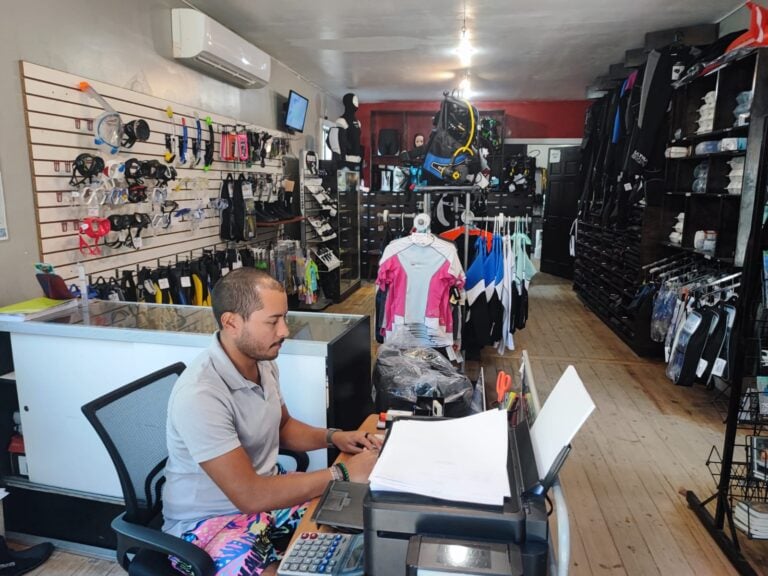 A man is working at a retail store in Roatan, Honduras, specializing in scuba gear and accessories with shelves displaying snorkel masks, fins, wetsuits, and diving equipment.