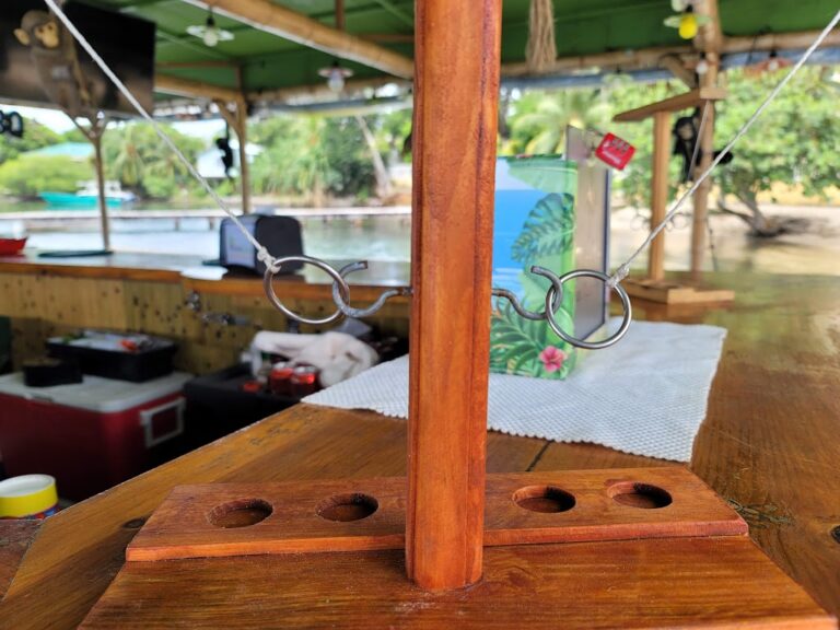 A wooden restaurant or bar counter with a metal ring and hook display stand on Roatan, Honduras, overlooking a river or lagoon with lush greenery and boats in the background.