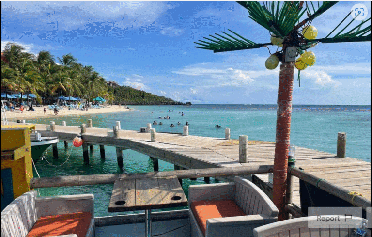 A scenic view of a wooden pier over clear turquoise water on Roatan, Honduras, with a beach lined with palm trees, colorful umbrellas, and people enjoying the ocean, seen from a seating area with cushioned chairs and a decorative palm tree with yellow ornaments.