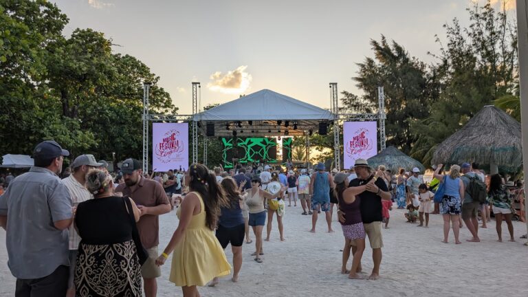 A lively outdoor music festival in Roatan, Honduras, features a stage with large screens displaying "Music Festival" signage, surrounded by groups of people dancing, socializing, and enjoying the event under a partly cloudy evening sky with lush green trees and tropical huts nearby.