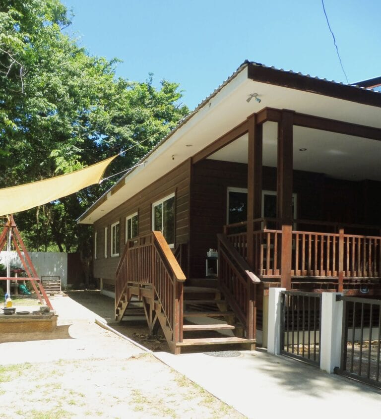 A two-story wooden house with a balcony and stairs, surrounded by lush green trees and a clear blue sky in Roatan, Honduras.